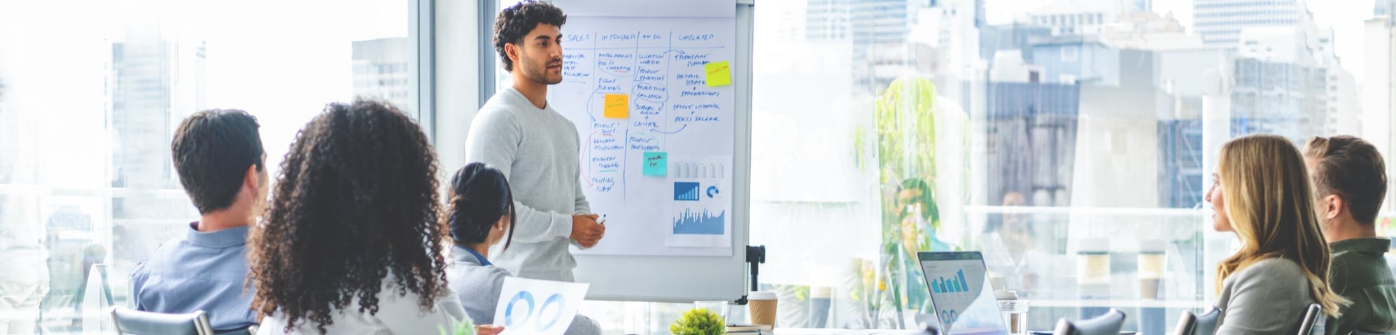 A group of business professionals in a modern conference room listening to a colleague give a presentation using a whiteboard with notes and charts, with city buildings visible through the large windows in the background.