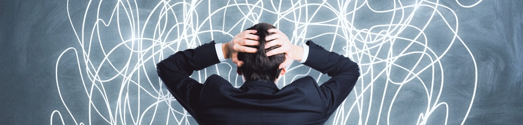 Frustrated businessman holding his head while facing a chalkboard filled with chaotic scribbles, symbolizing confusion and disorganization