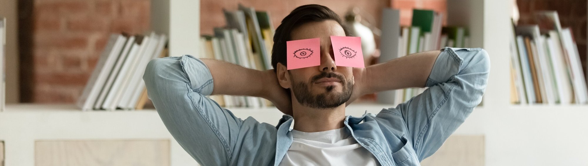 Man relaxing at work with sticky notes with eyes drawn on them on his face [1338118940]_banner