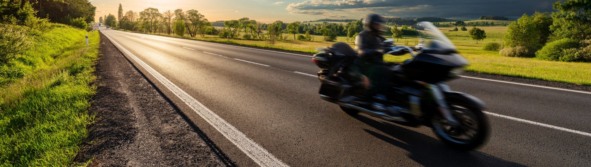 Motion blurred black motorcycle riding on an empty asphalt road in a rural landscape at sunset [1324281558]_banner-1