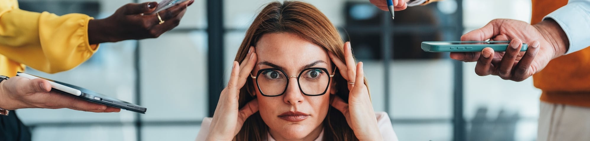 A stressed woman in glasses holds her head in her hands while multiple people reach phones and tablets toward her from all directions