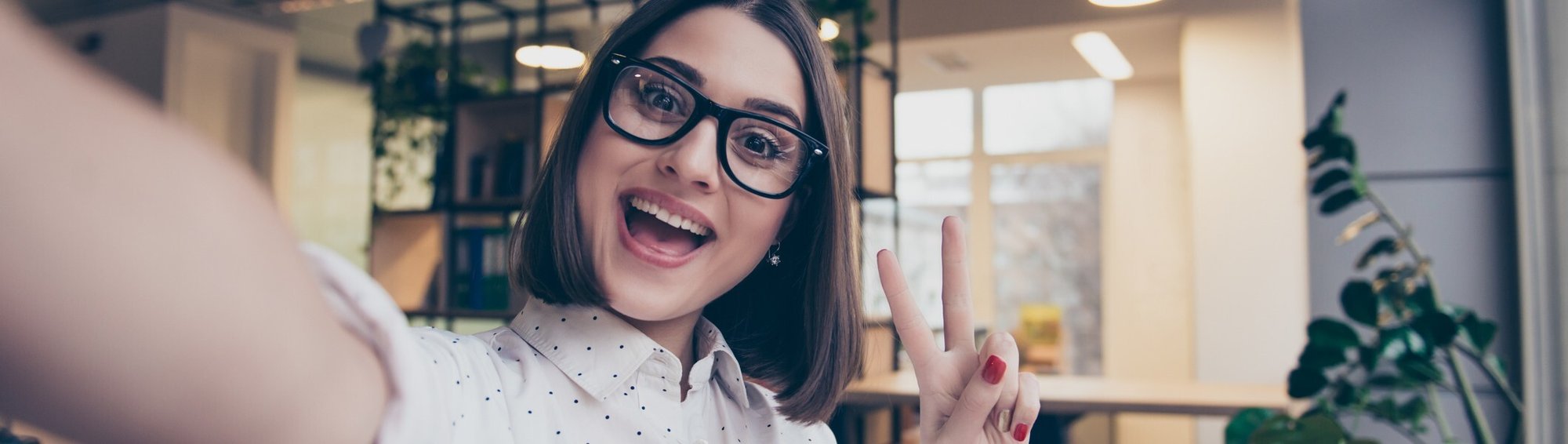 Smiling young woman wearing glasses takes a cheerful selfie indoors, flashing a peace sign with her hand. Smiling young woman wearing glasses takes a cheerful selfie indoors, flashing a peace sign with her hand.